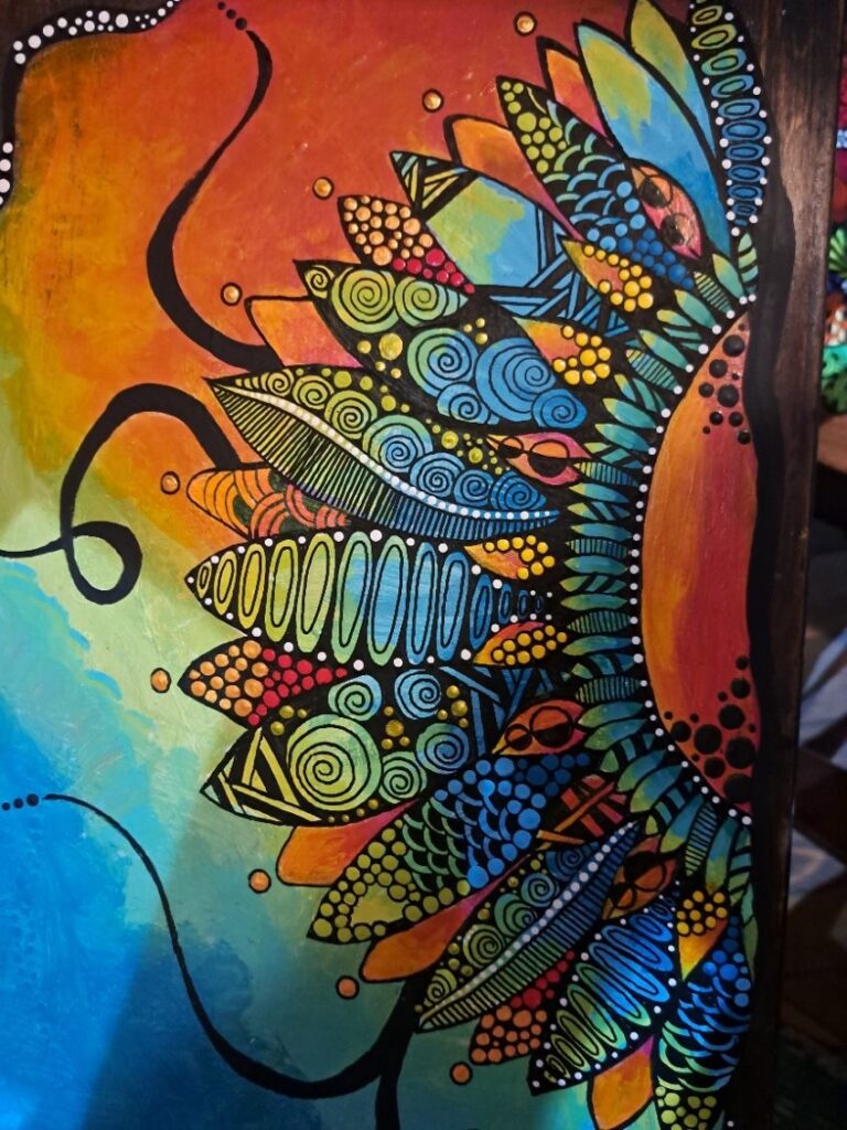 Vibrant folkedelic artwork with layered, petal-like shapes filled with spirals, dots, and geometric patterns, set against a warm-to-cool gradient background blending orange, yellow, blue, and green.