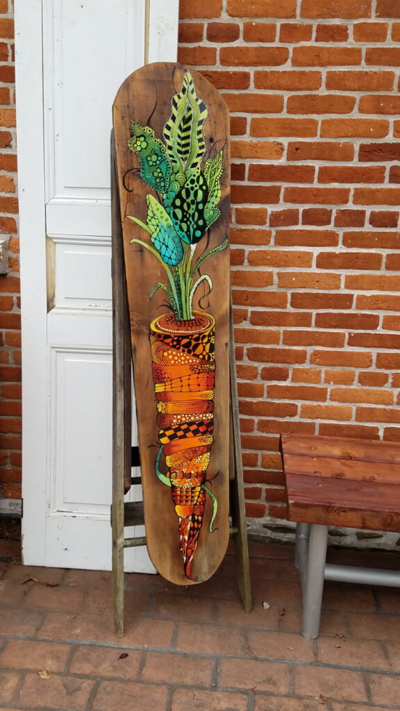 Vibrant folkedelic artwork featuring a stylized carrot with layered, leaf-like shapes filled with spirals, dots, and geometric patterns, painted on a vertical wooden board.