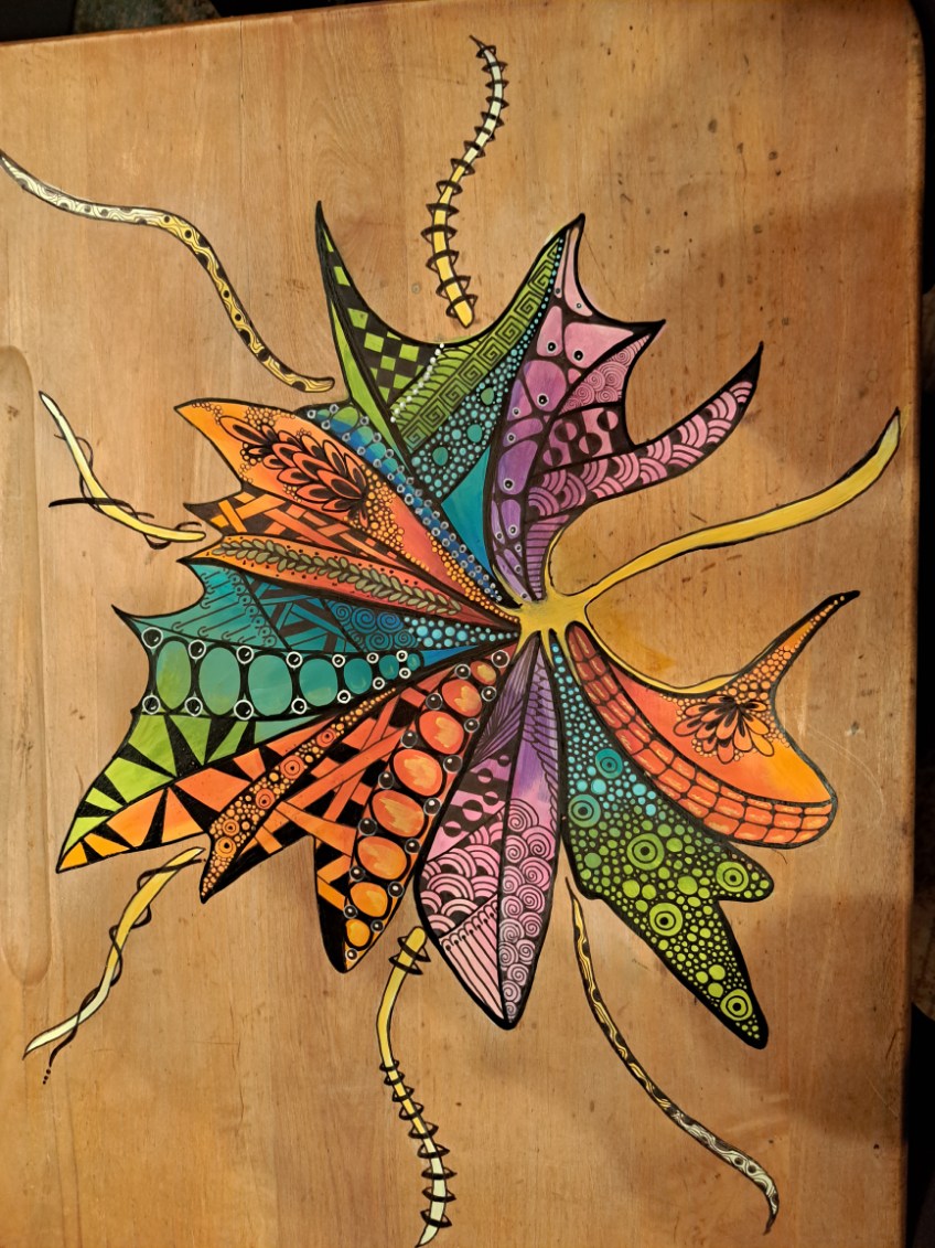 Colorful folkedelic leaf design composed of segmented, petal-like shapes filled with intricate patterns, including spirals, checkerboards, and dots, painted on a warm-toned wooden surface.