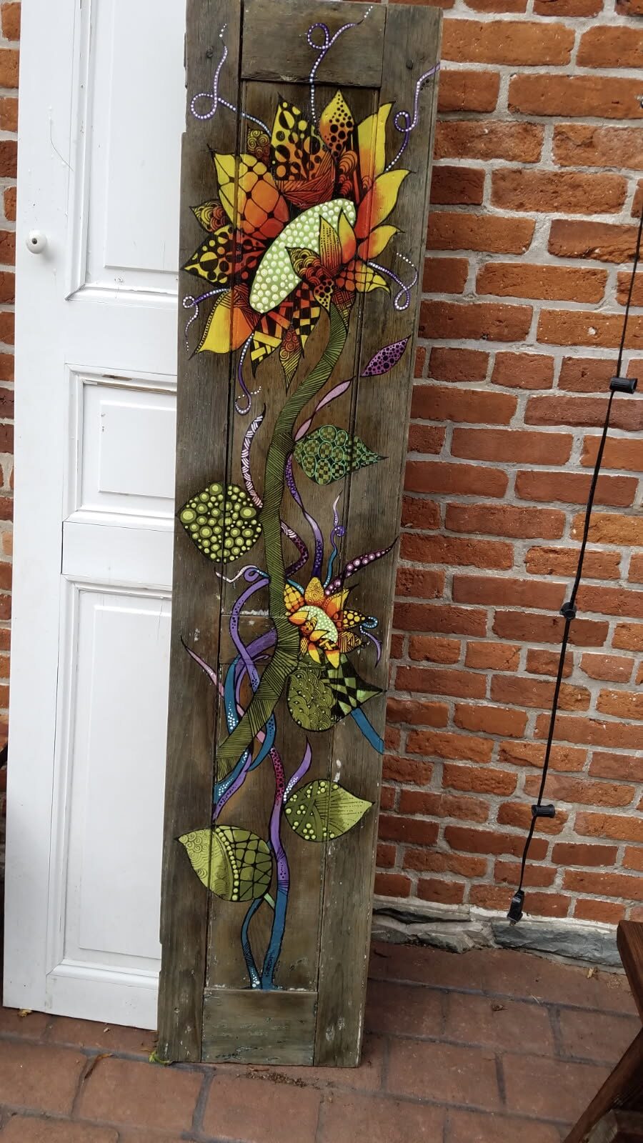 Colorful folkedelic floral design with intricate, patterned petals in warm tones of orange and yellow, connected by a flowing green stem with decorative leaves, painted on a rustic wooden panel.