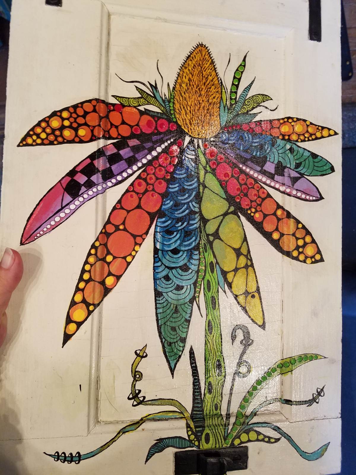 Colorful folkedelic flower design with long, patterned petals in bold hues of orange, red, blue, green, and purple, filled with dots, checkerboards, and swirling motifs on a painted surface.